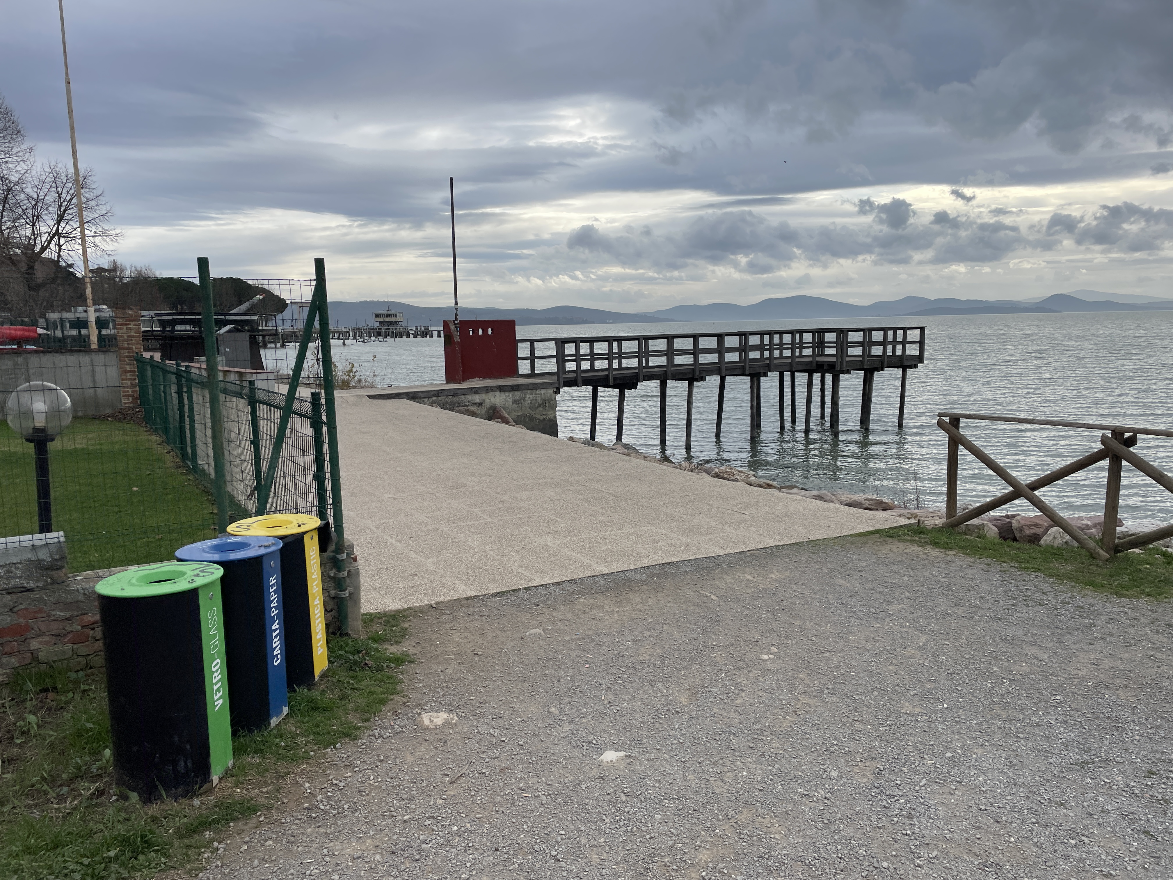 Access to lake with wooden pier. On left green metal fence and trash cans. At right an interrupted fence.